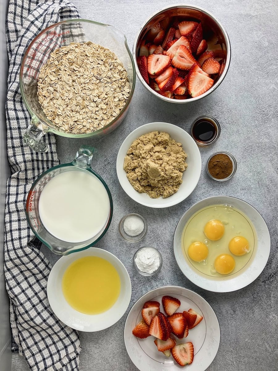Ingredients for making strawberry baked oatmeal in prep bowls. Rolled oats, sliced strawberries, brown sugar, eggs, milk, salt, cinnamon, baking powder, vanilla, and melted butter.