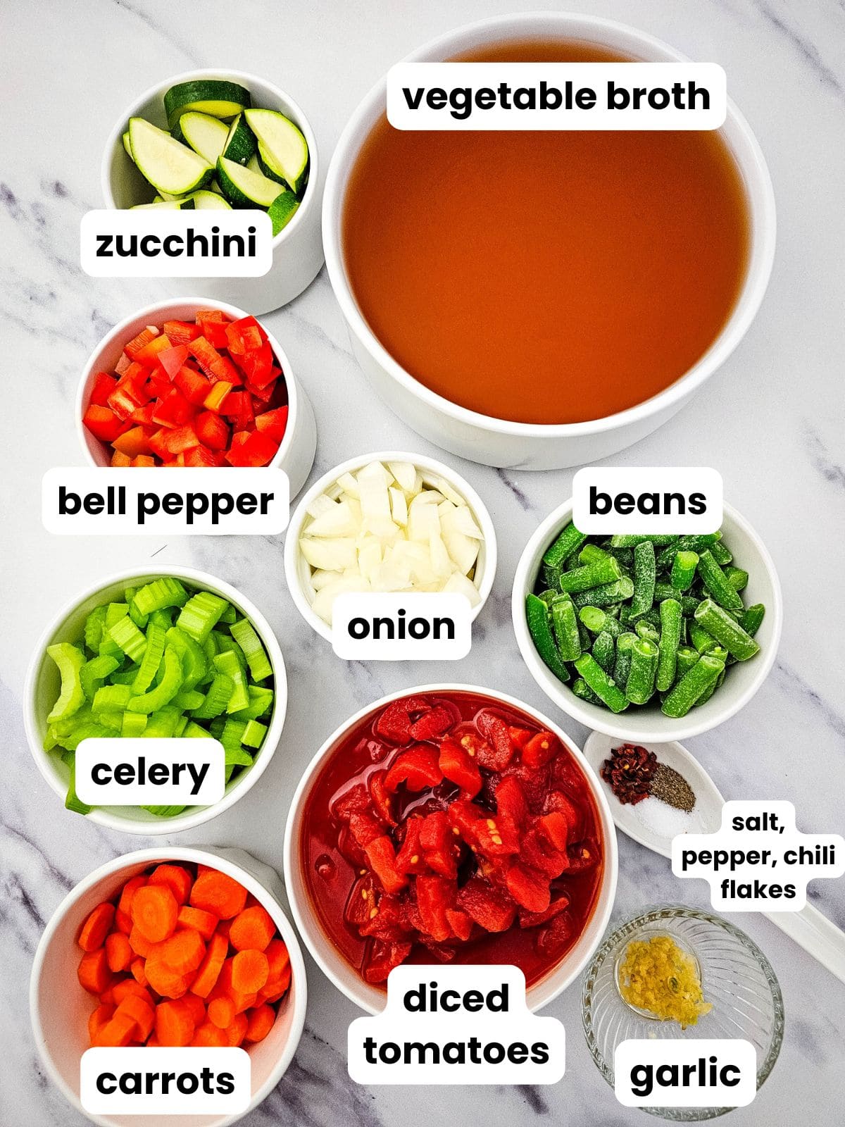 Ingredients for the vegetable soup in bowls including vegetable broth, diced tomatoes, zucchini, red bell pepper, onion, green beans, celery, carrots, garlic, salt, pepper, and chili flakes.
