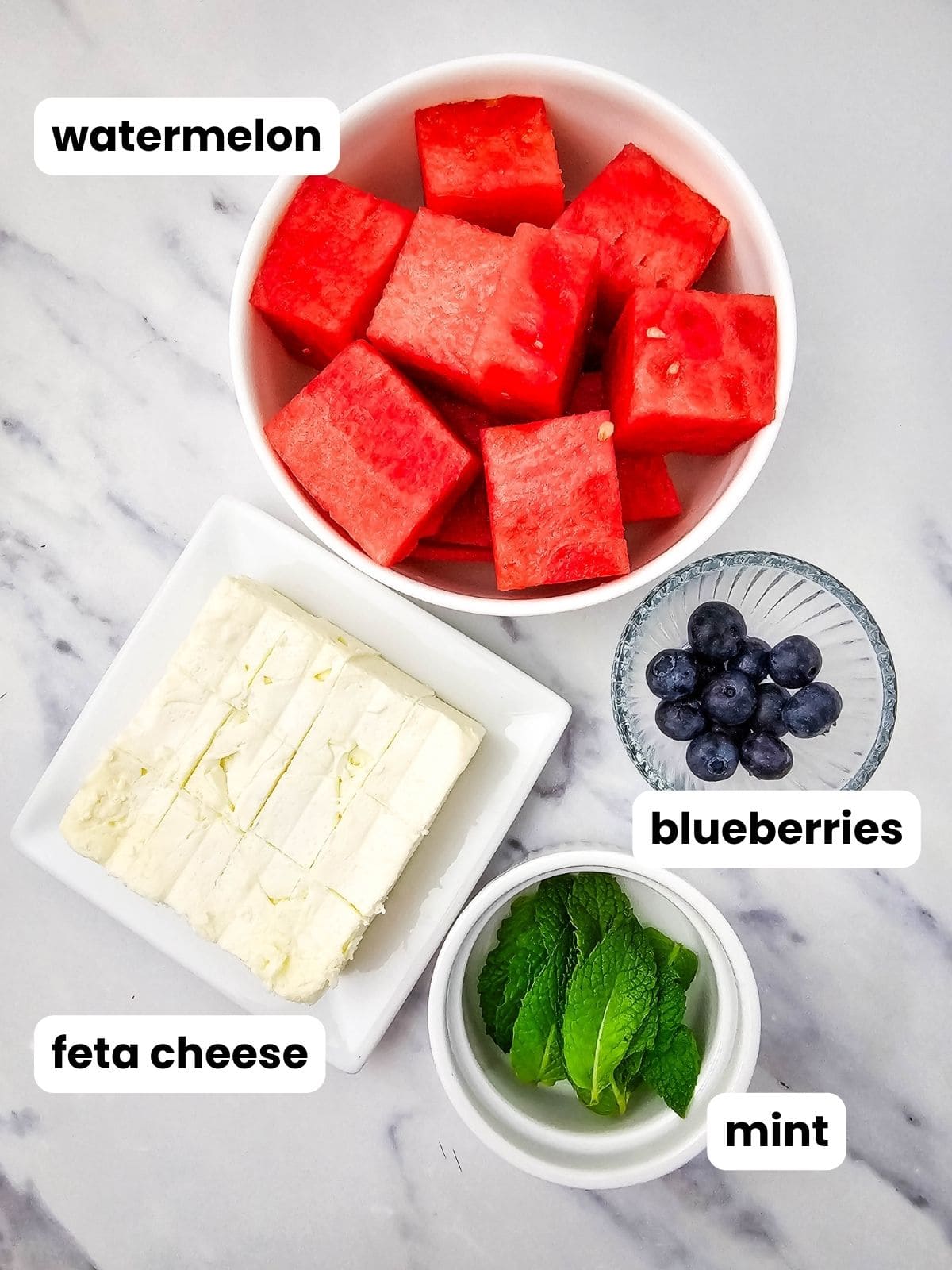 Skewer ingredients in a bowl of cubed watermelon, cubed feta cheese, blueberries, and fresh mint leaves.
