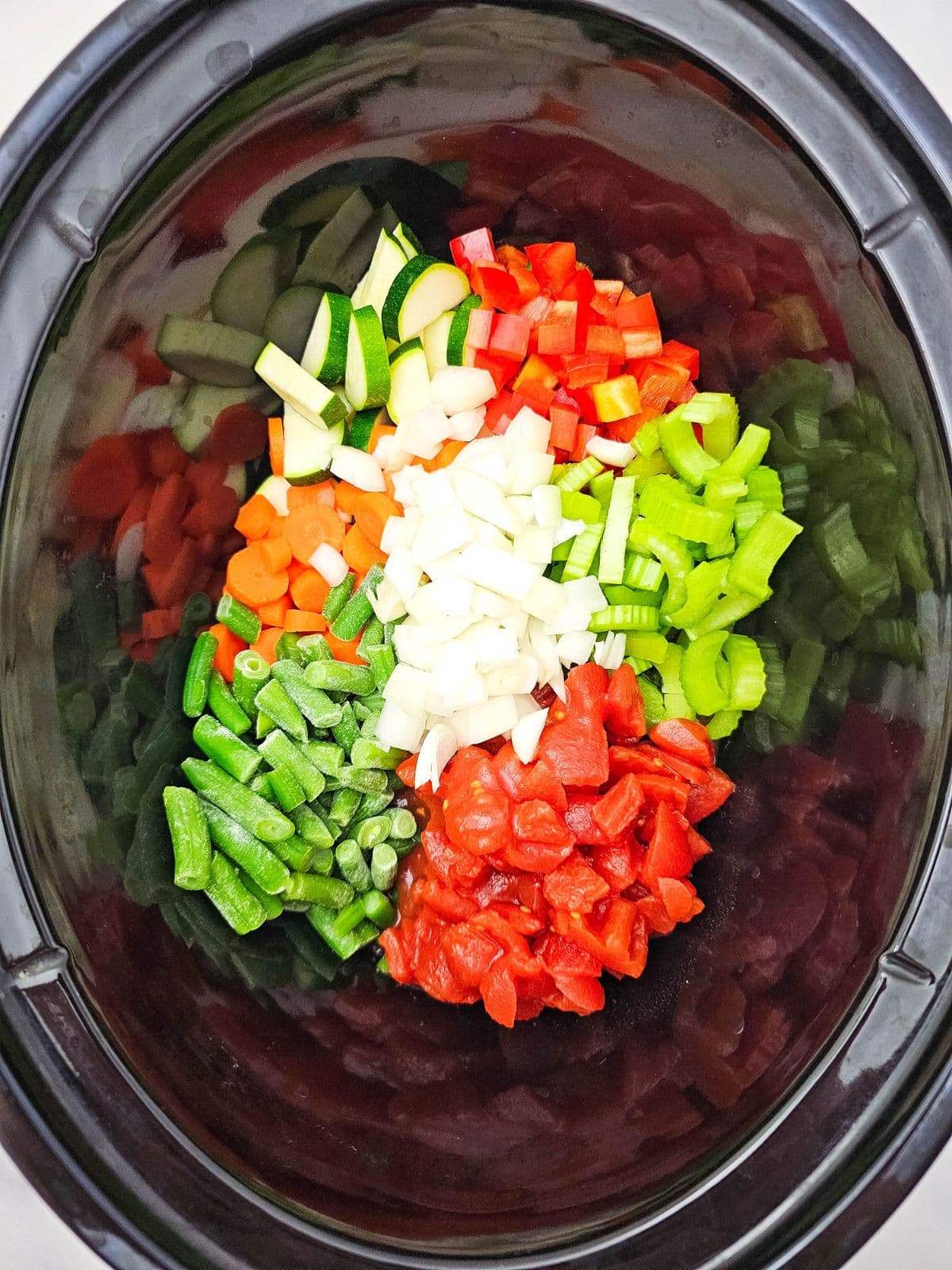 Sliced and diced vegetables in a slow cooker, including green beans, peppers, celery, carrots, zucchini, onions, and diced tomatoes.