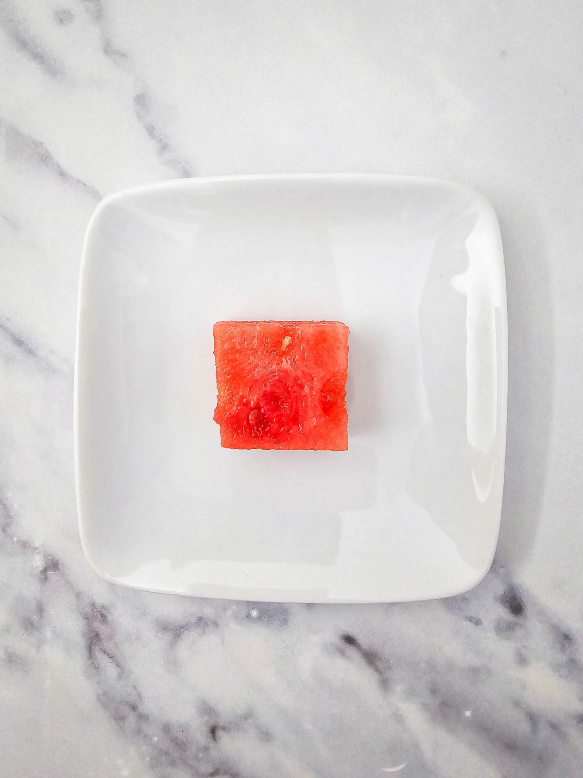 A square cube of watermelon on a white plate.