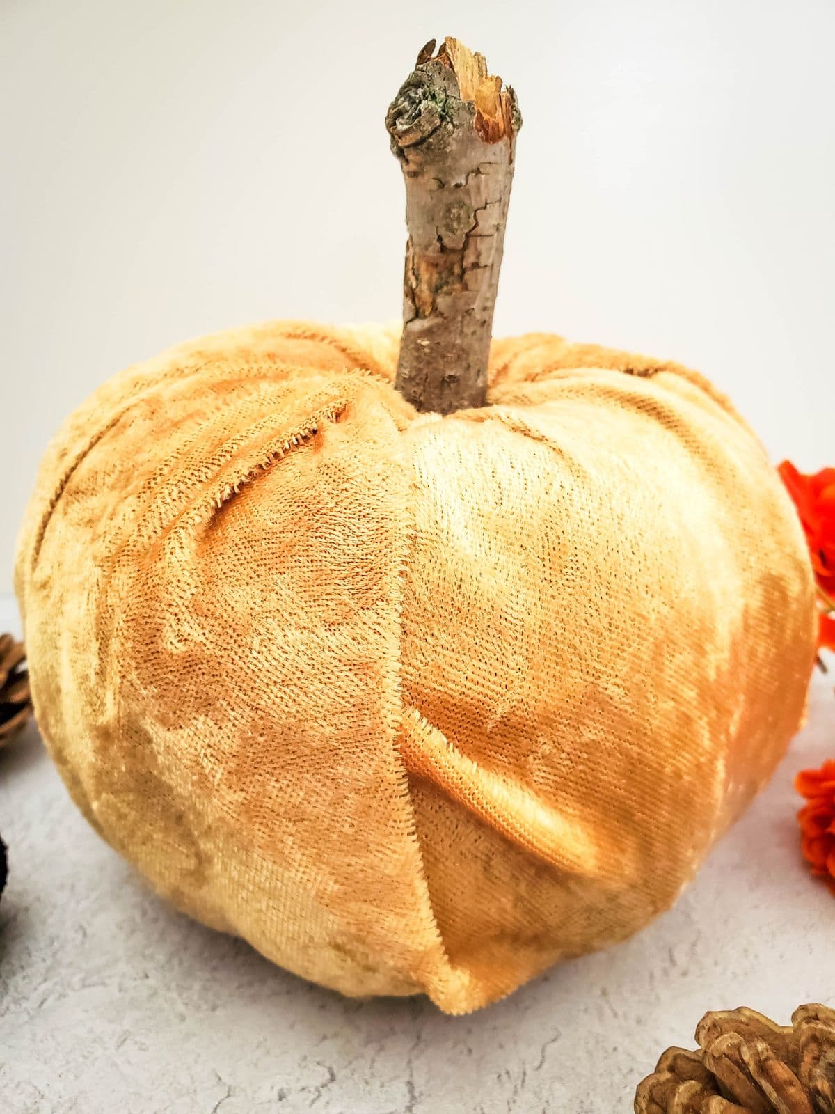 A no sew velvet pumpkin with a stick stem sitting on the counter next to orange flowers and pinecones.