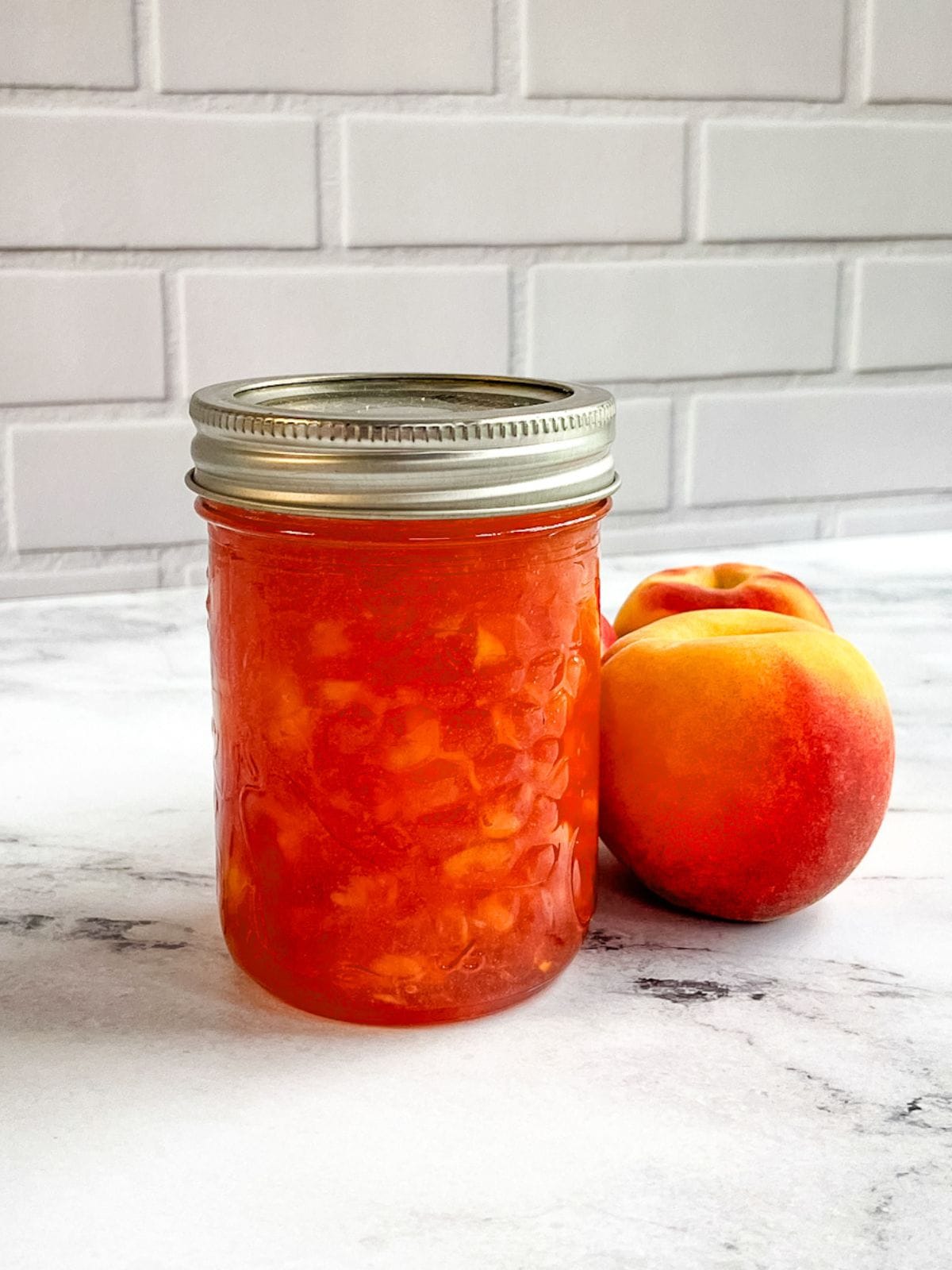 A jar of homemade peach jam is on the counter next to fresh peaches.