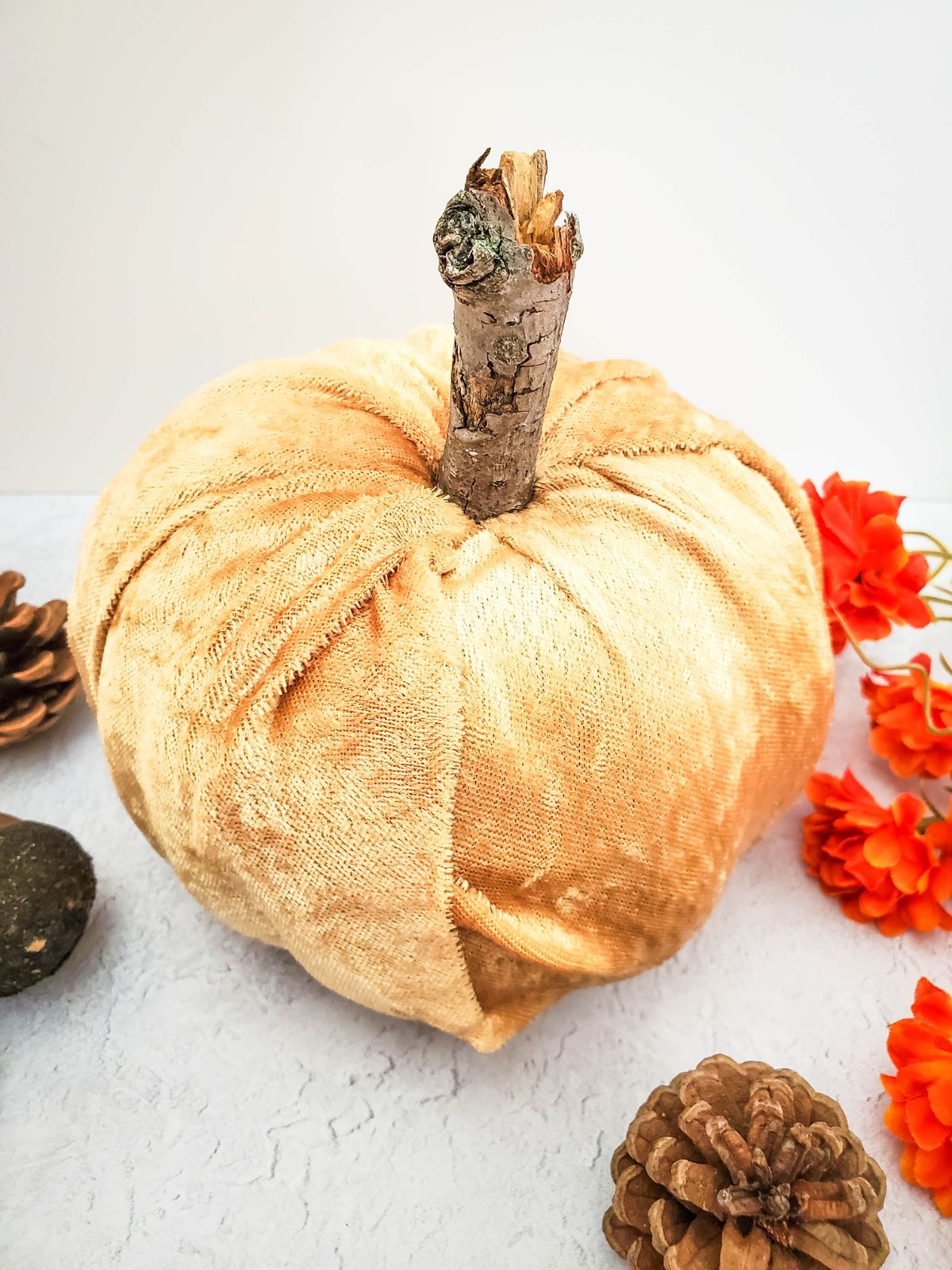 A DIY no sew velvet pumpkin sitting on the counter next to pinecones and orange flowers.