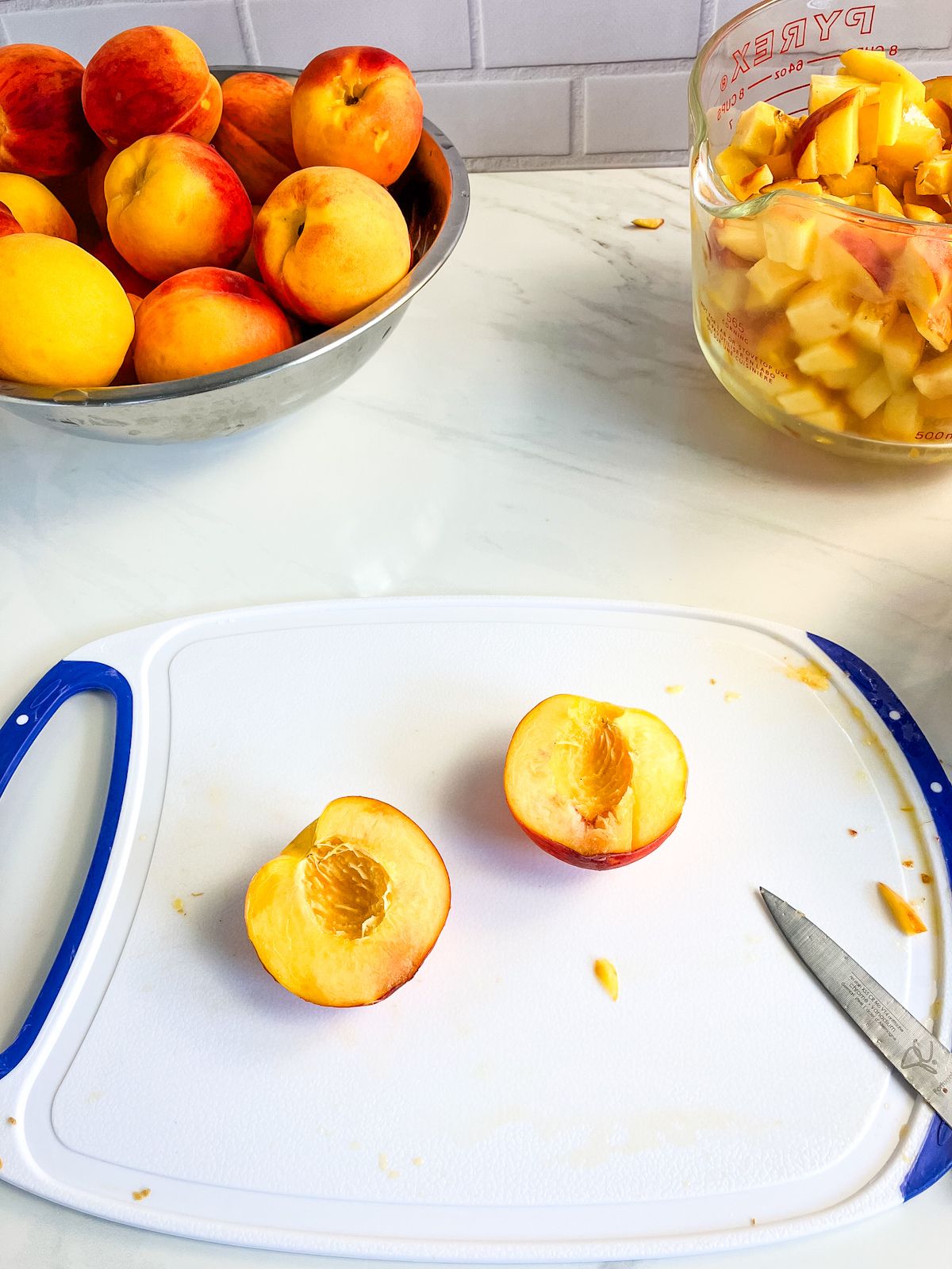 A bowl of peaches, a peach cut in half on a cutting board, and a measuring cup of diced peaches.