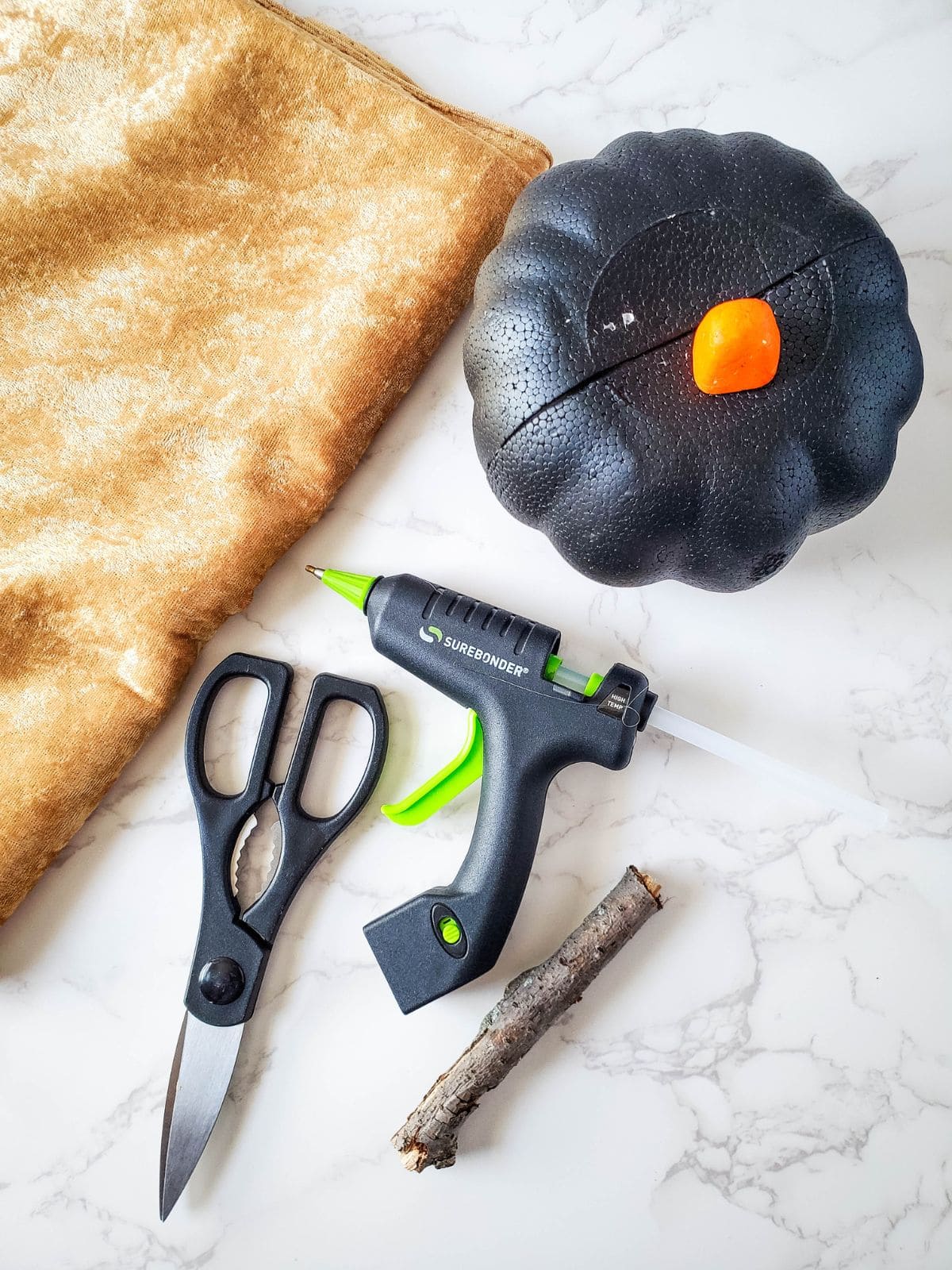 Supplies for making a velvet pumpkin: velvet cloth, a foam pumpkin, a glue gun with glue, scissors, and a stick.