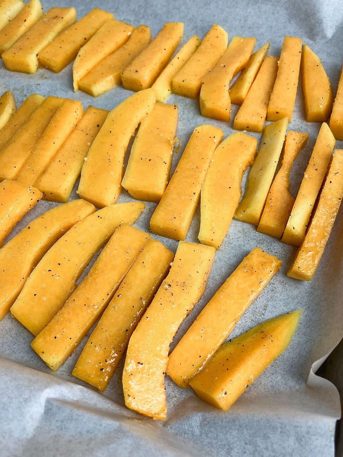 Seasoned raw butternut squash fries are spread out on a parchment-lined baking sheet.