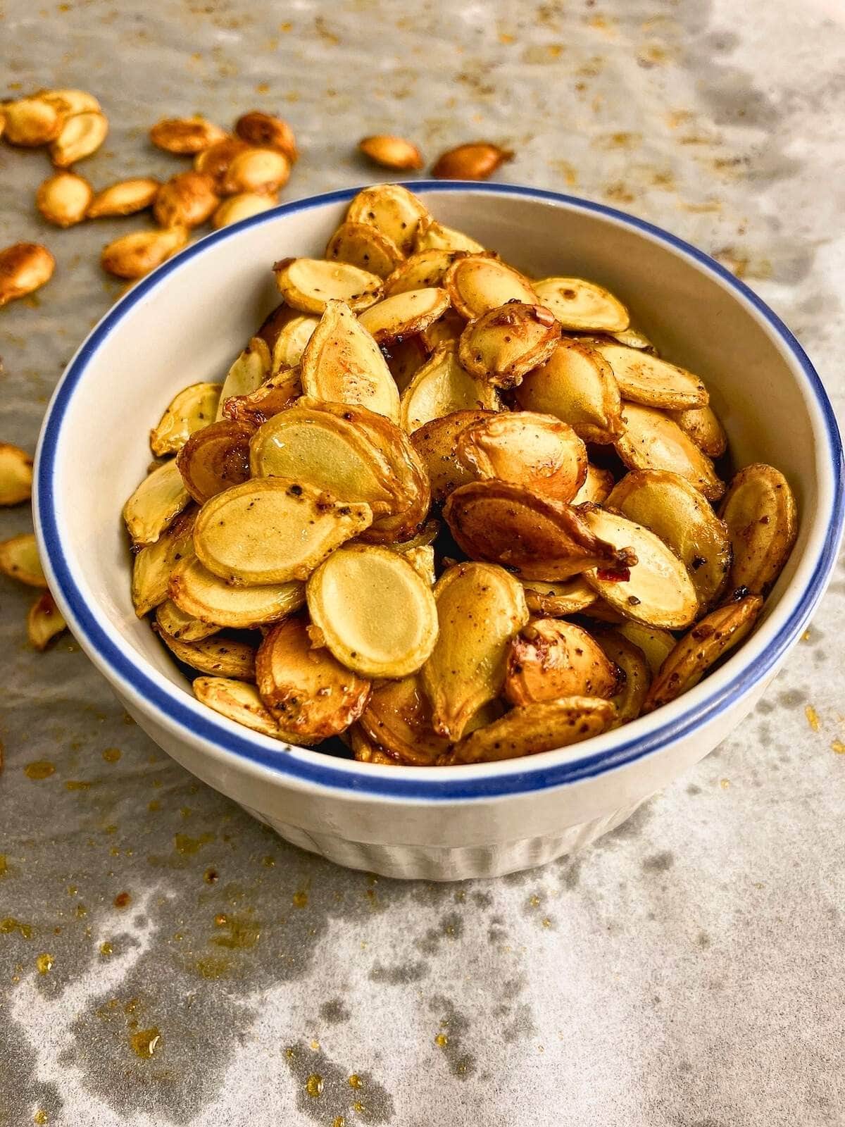 Spicy roasted pumpkin seeds in a small white bowl with some still on the pan next to it.