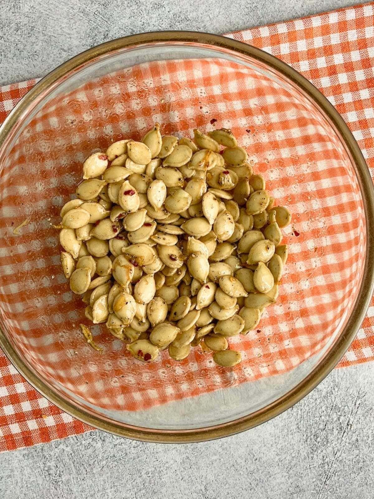 Raw pumpkin seeds are tossed in a mixing bowl with vegetable oil and spicy seasonings.