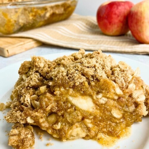 A slice of homemade apple pumpkin crisp on a white plate with a baking dish of crisp and fresh apples in the background.