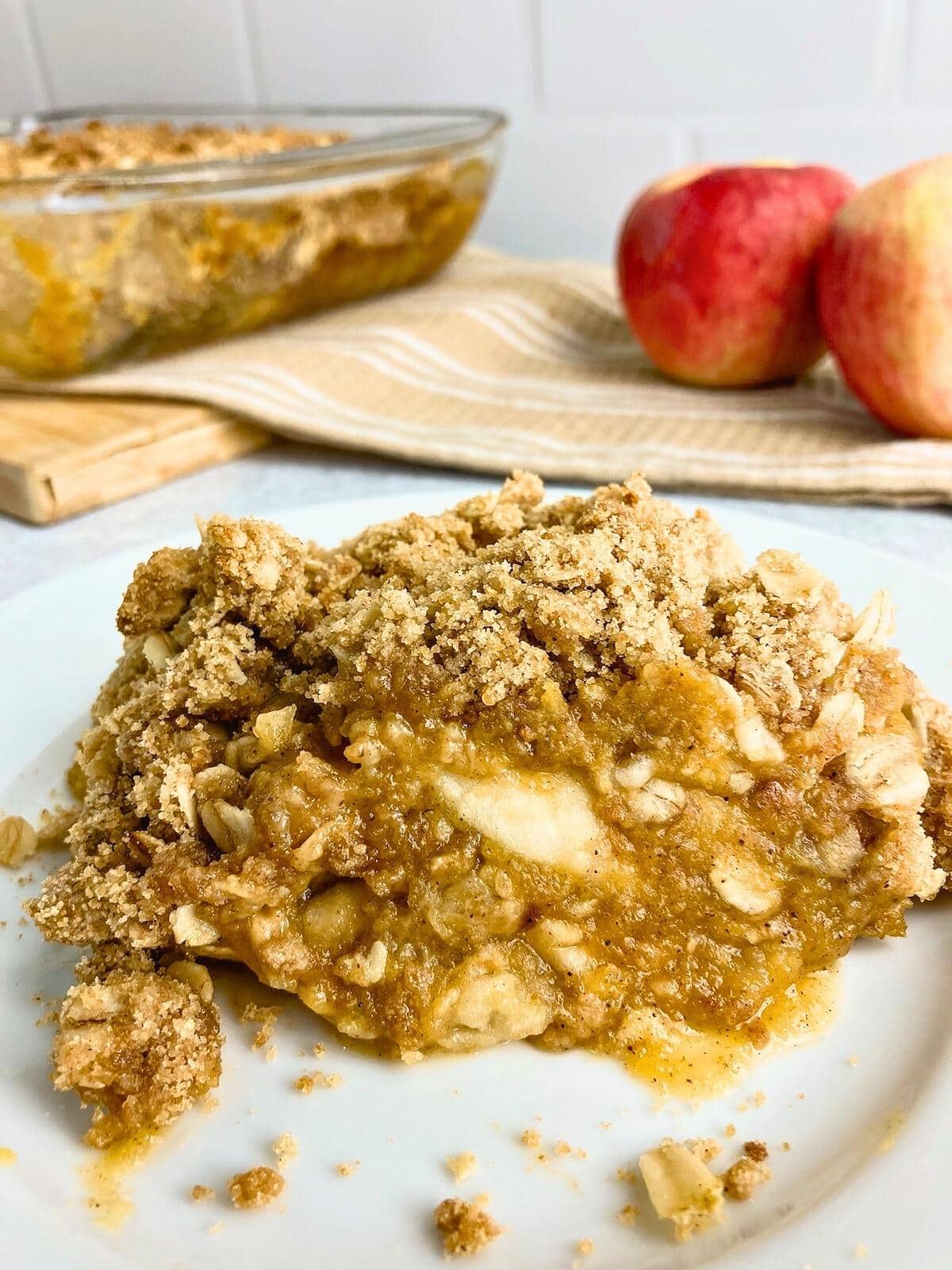 A slice of homemade apple pumpkin crisp on a white plate with a baking dish of crisp and fresh apples in the background.