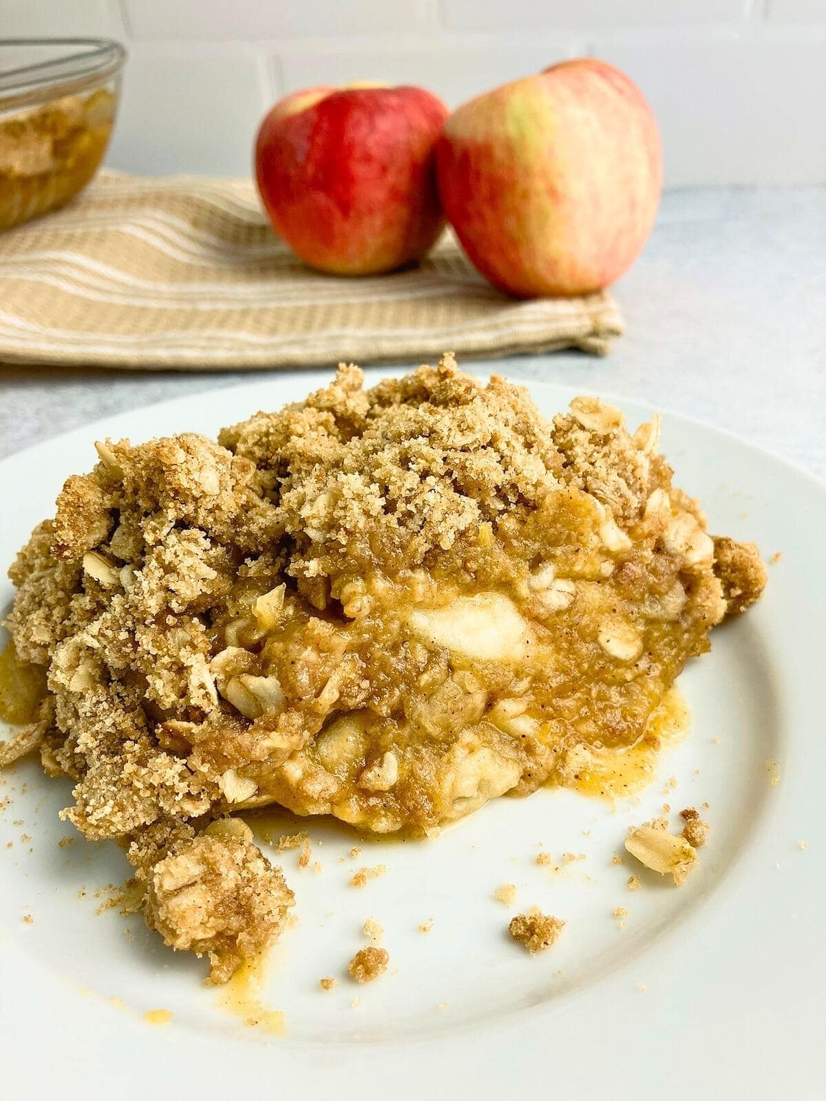A slice of homemade apple pumpkin crisp on a white plate with fresh apples in the background.