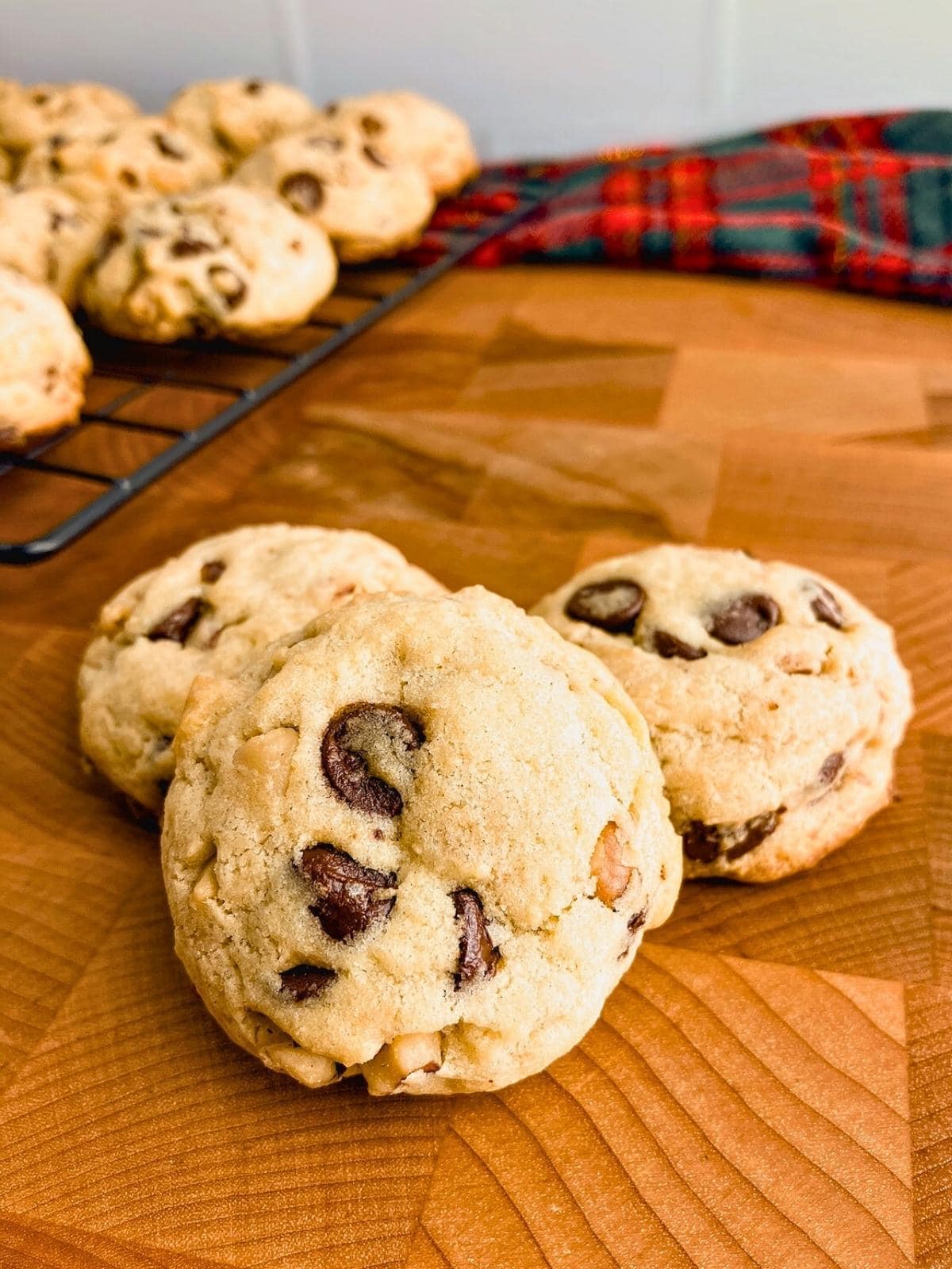Three walnut chocolate chip cookies on a wooden table next to a cooking rack of homemade cookies.