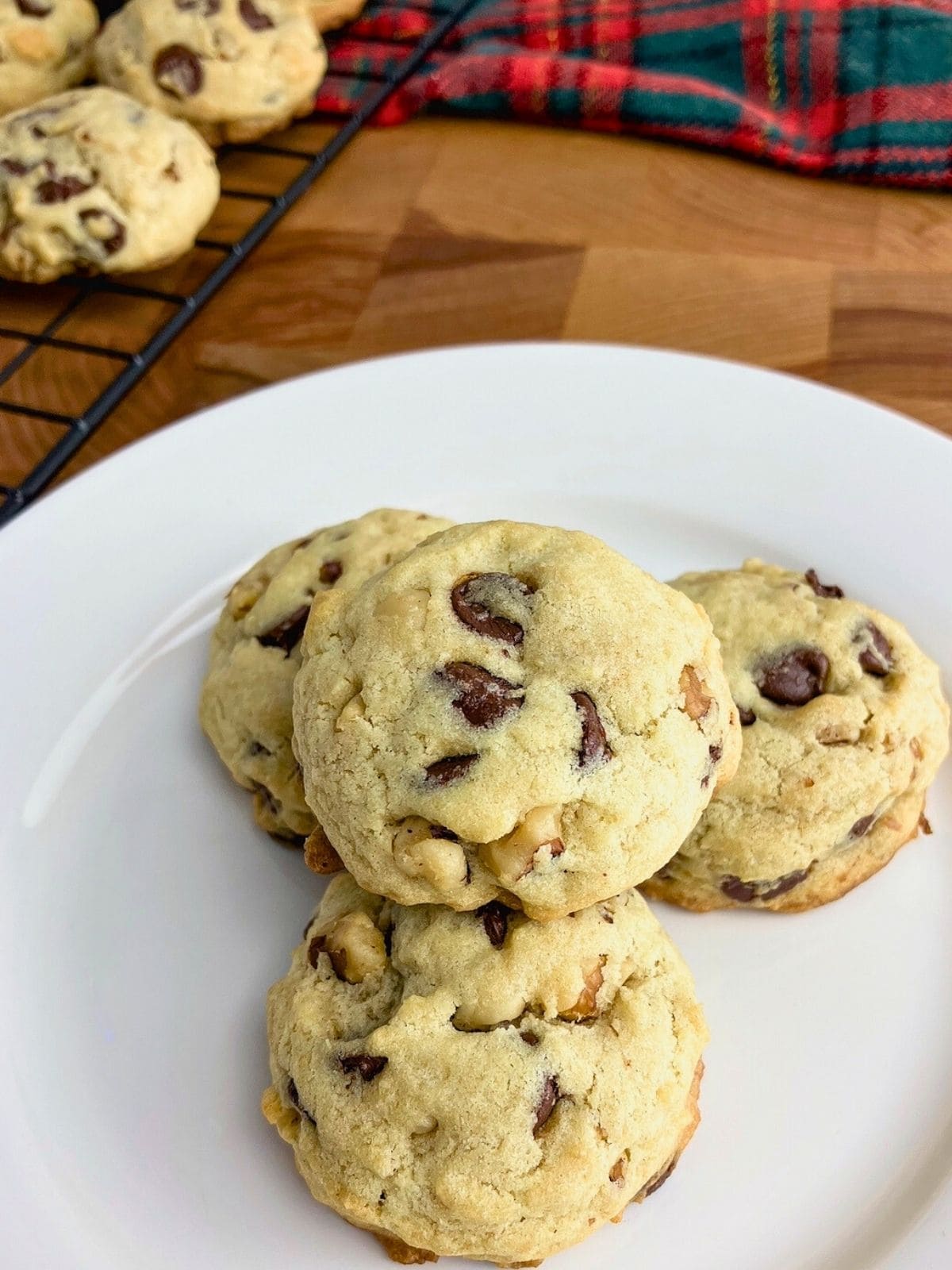 Four chocolate chip walnut cookies on a white plate next to a cooling rack of cookies.