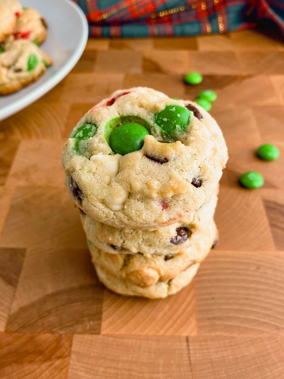 A stack of chocolate chip cookies with M&Ms on a wooden table next to a platter of cookies.