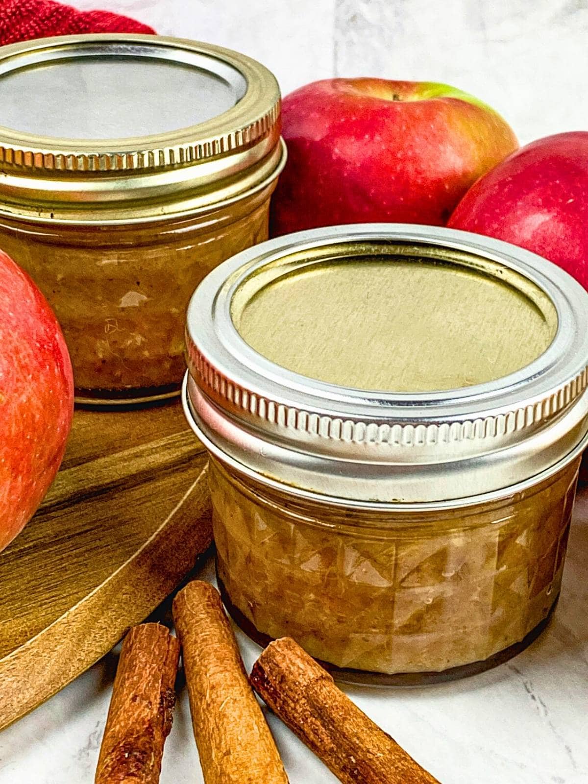 Two jars of homemade apple chutney next to fresh apples and cinnamon sticks.