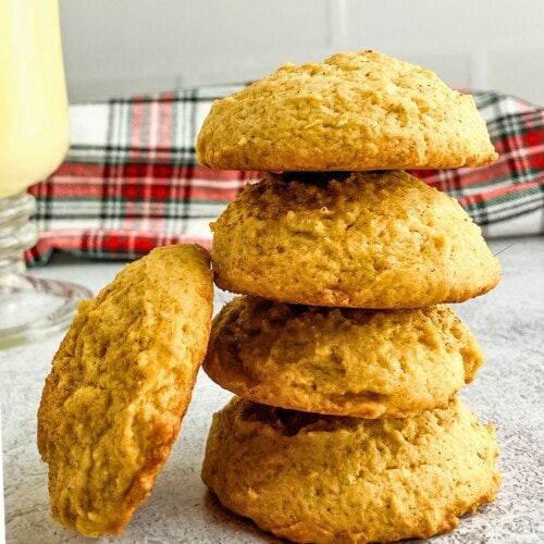 Soft eggnog cookies stacked on the counter next to a glass of eggnog.