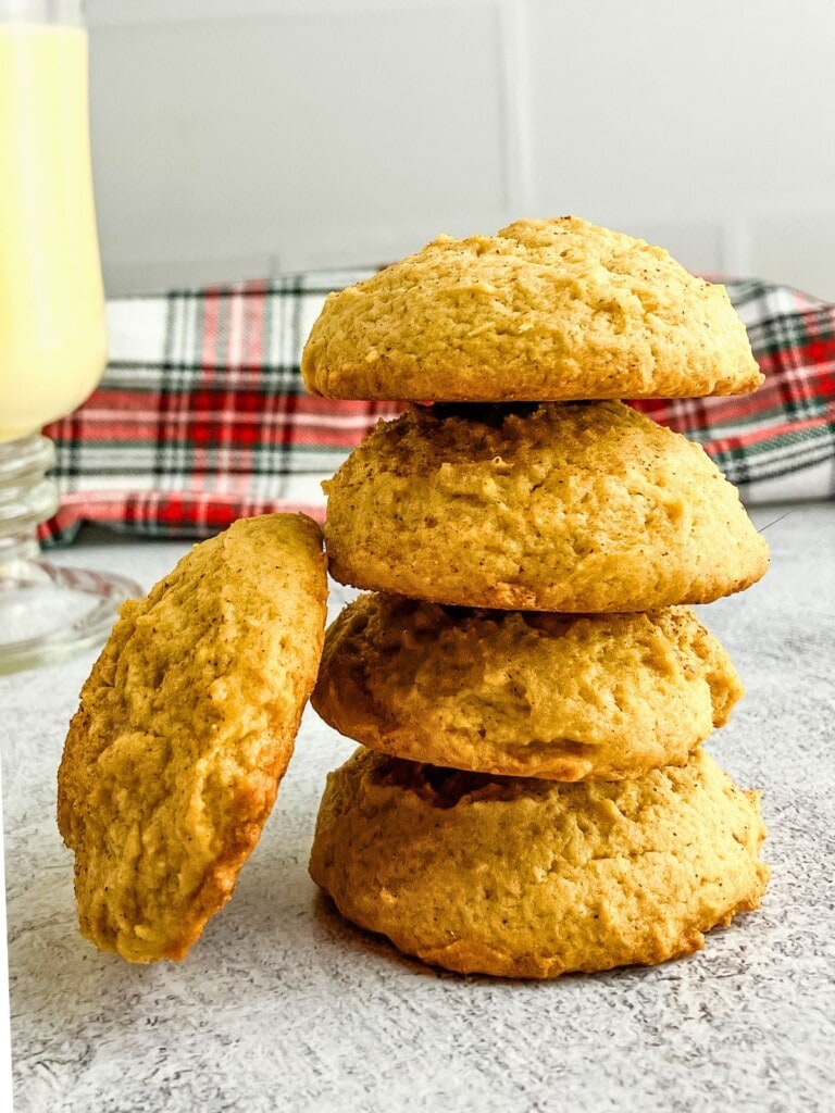 Soft eggnog cookies stacked on the counter next to a glass of eggnog.
