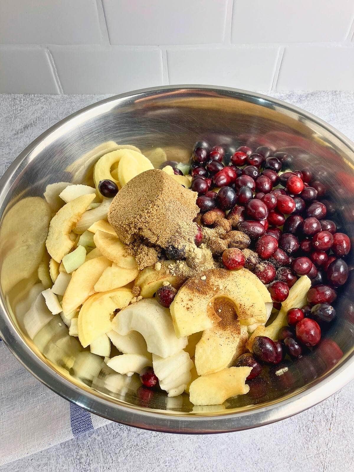 A large mixing bowl filled with apple slices, cranberries, brown sugar, and spices.