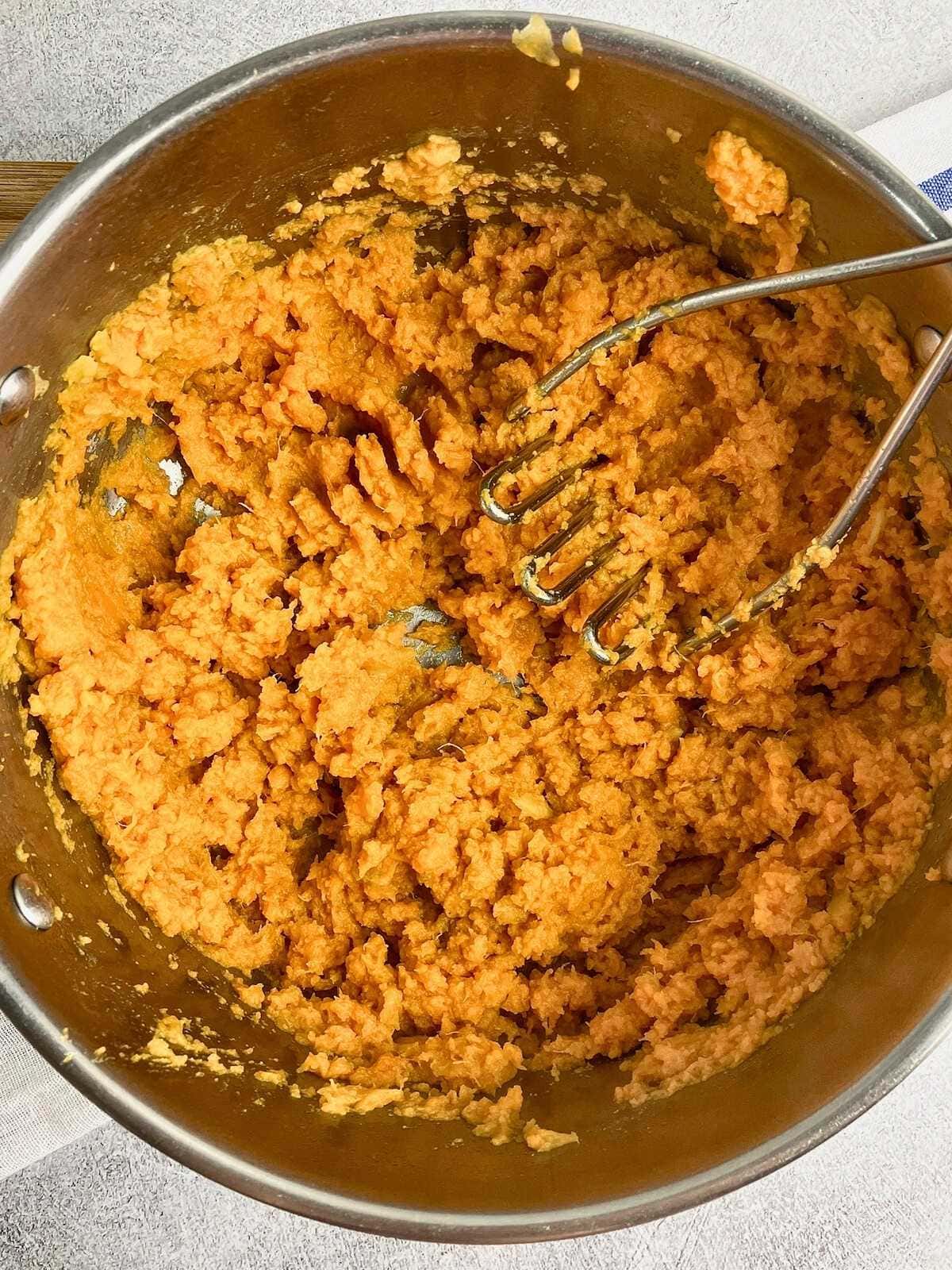 Sweet potatoes are mashed in a pot with the potato masher leaning against the side of the pot.