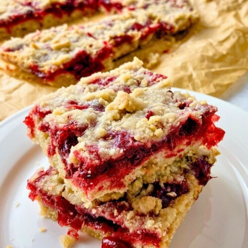 Two cranberry crumb bars stacked on a plate next to a sheet of parchment with more cranberry squares.
