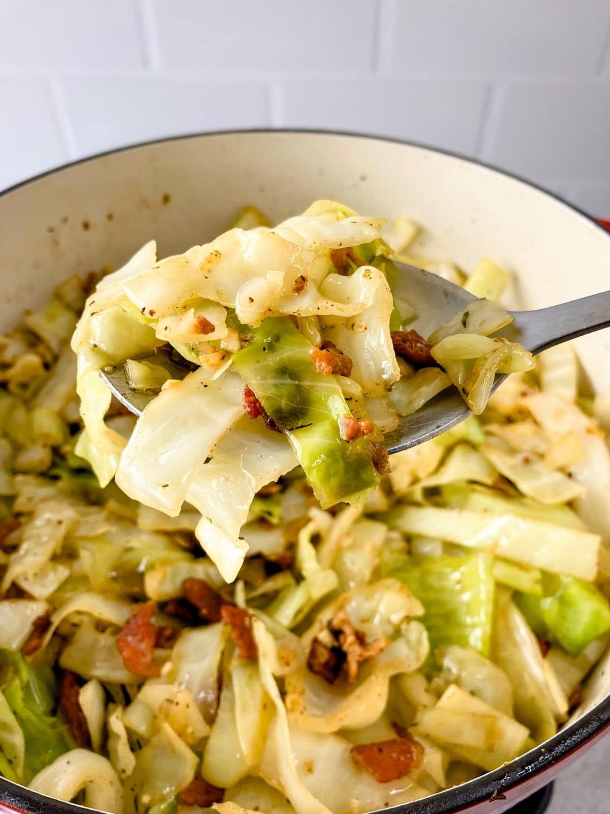A large cast iron Dutch oven filled with homemade fried cabbage and bacon with a serving spoonful lifted up.