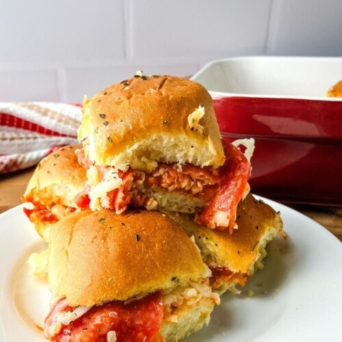 Pizza sliders on Hawaiian rolls are stacked on a plate next to a baking dish.