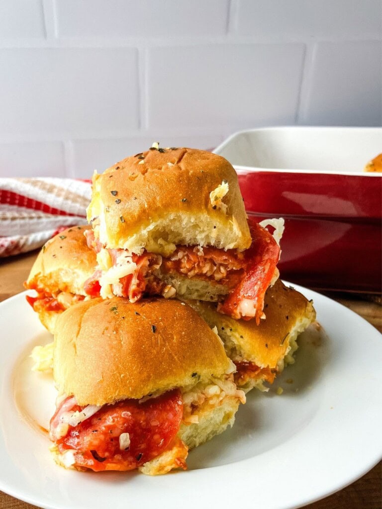 Pizza sliders on Hawaiian rolls are stacked on a plate next to a baking dish.