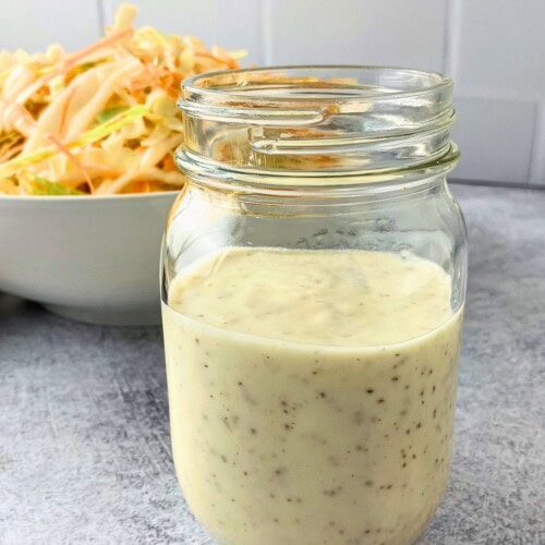 Creamy coleslaw dressing in a jar on the counter next to a bowl of homemade slaw.