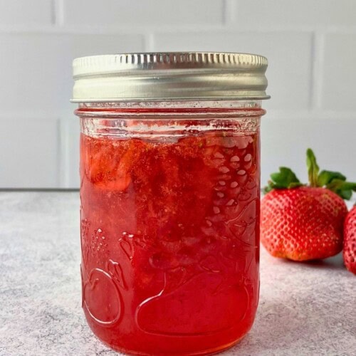 A jar of classic homemade strawberry jam is sitting on the counter next to fresh strawberries.
