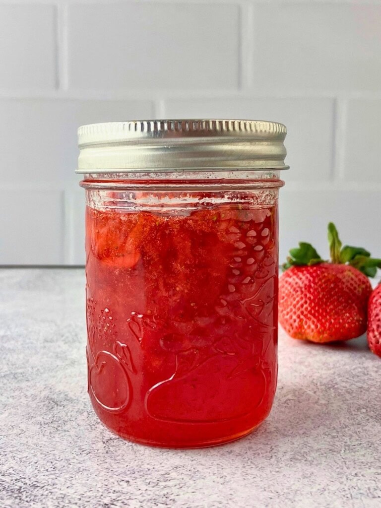 A jar of classic homemade strawberry jam is sitting on the counter next to fresh strawberries.