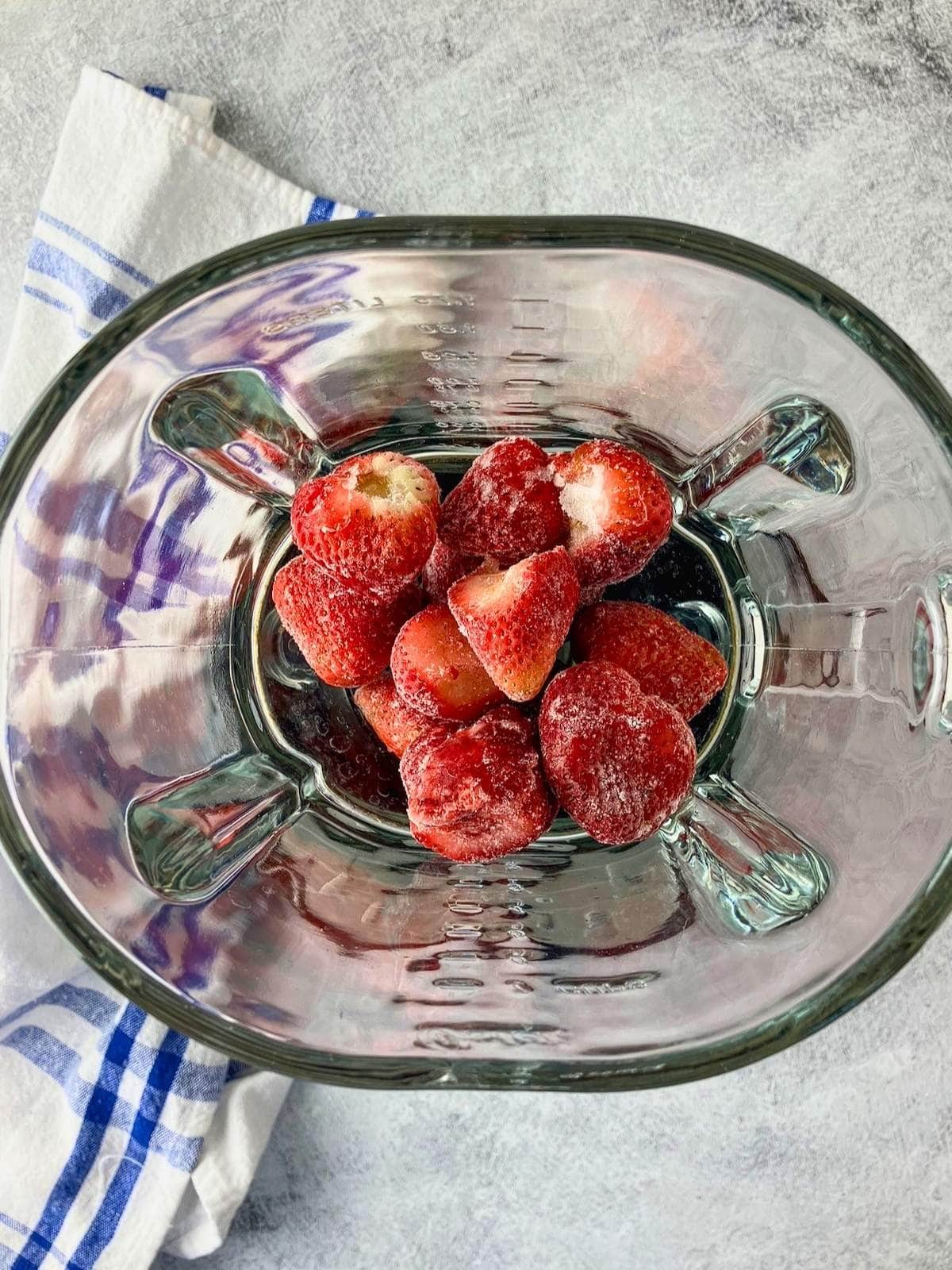 Frozen strawberries sit in the bottom of a glass blender. A blue and white towel rests beside the blender on a gray countertop.