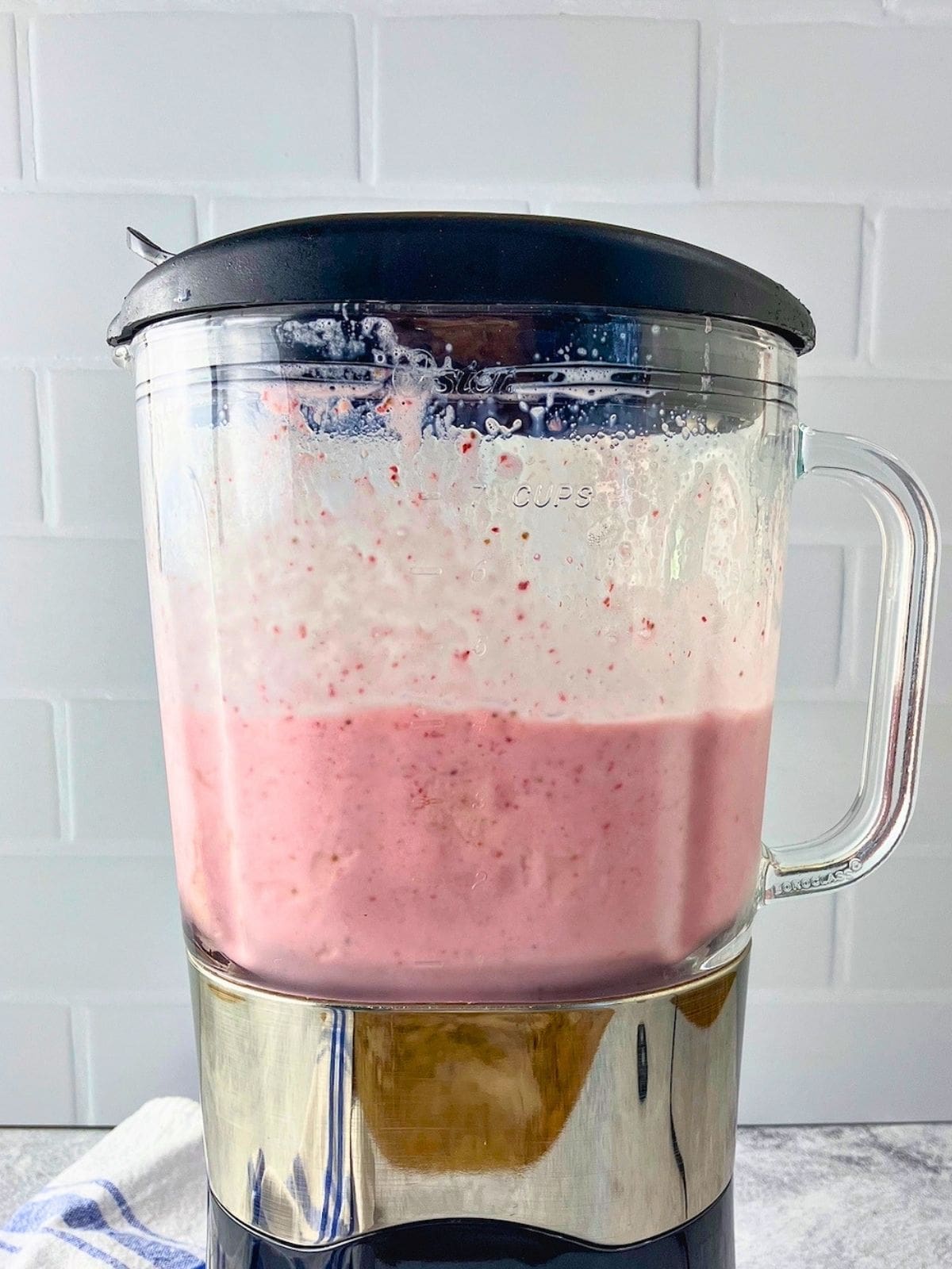 Pink strawberry milk blended in a glass blender with the lid on. Splashes of the drink mark the sides of the blender against a white tile backsplash.