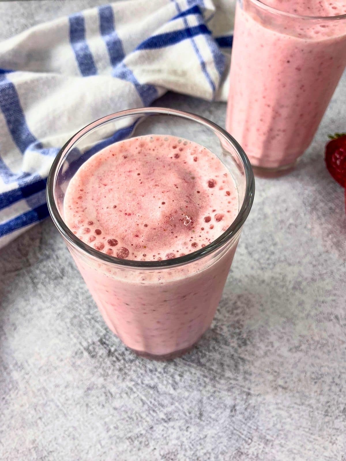 Two glasses of pink strawberry milk with real strawberries blended in. A whole strawberry sits beside the back glass on a gray countertop with a blue and white striped towel.