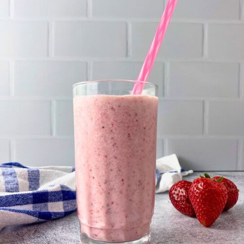 A tall glass of strawberry milk with real strawberries blended in and a pink straw. Fresh strawberries and a blue and white towel sit beside it on a gray countertop.