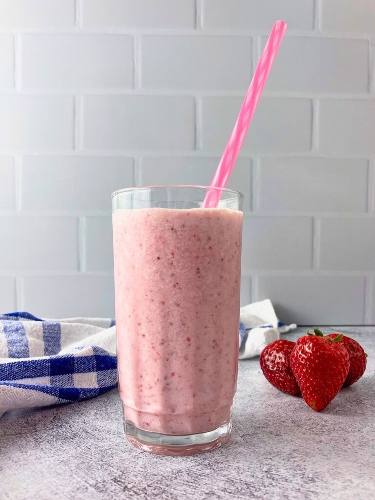 A tall glass of strawberry milk with real strawberries blended in and a pink straw. Fresh strawberries and a blue and white towel sit beside it on a gray countertop.
