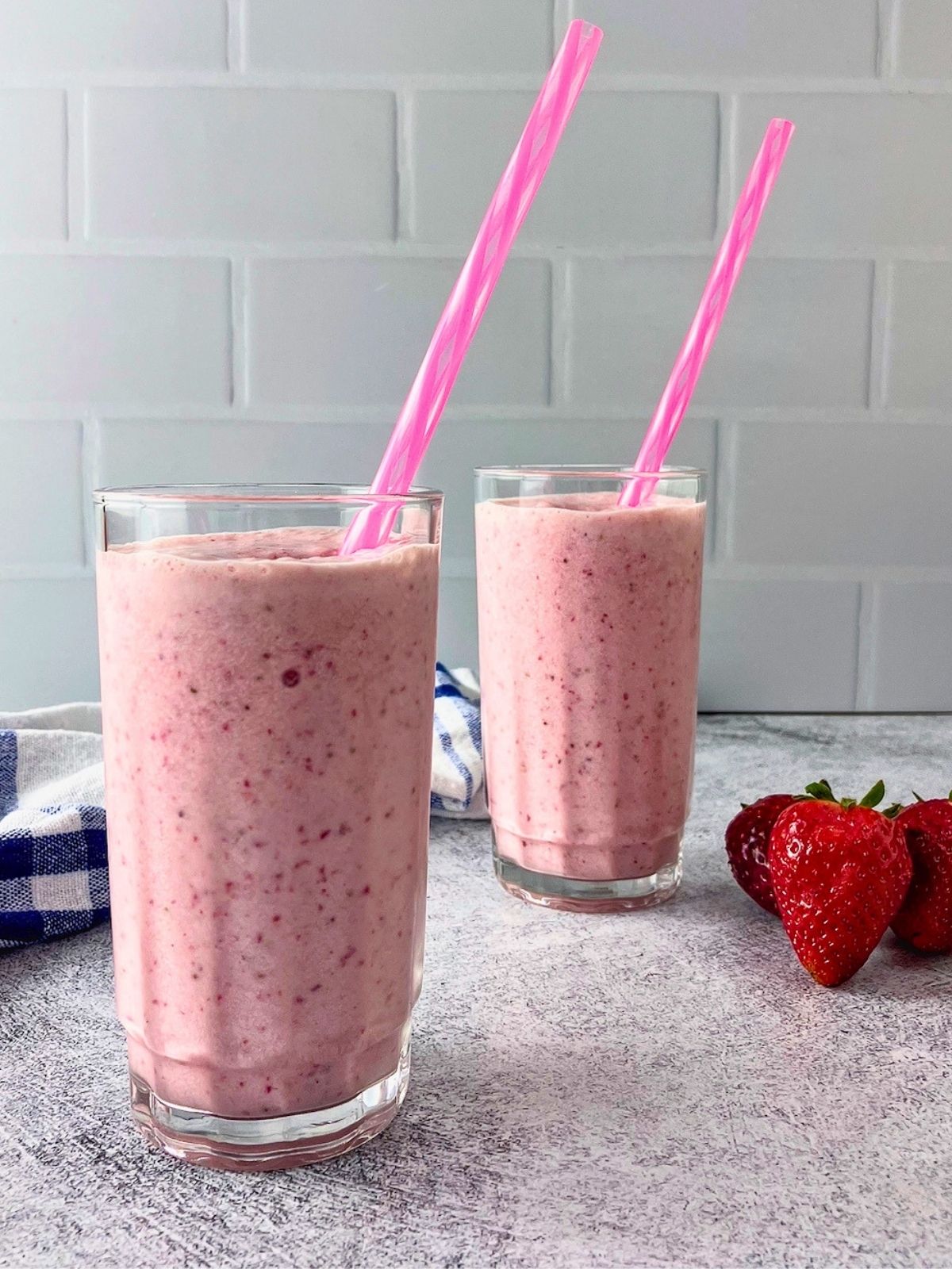 Two glasses of pink strawberry milk with real strawberries blended in and pink straws. Fresh strawberries and a blue and white towel sit on the gray countertop beside them.