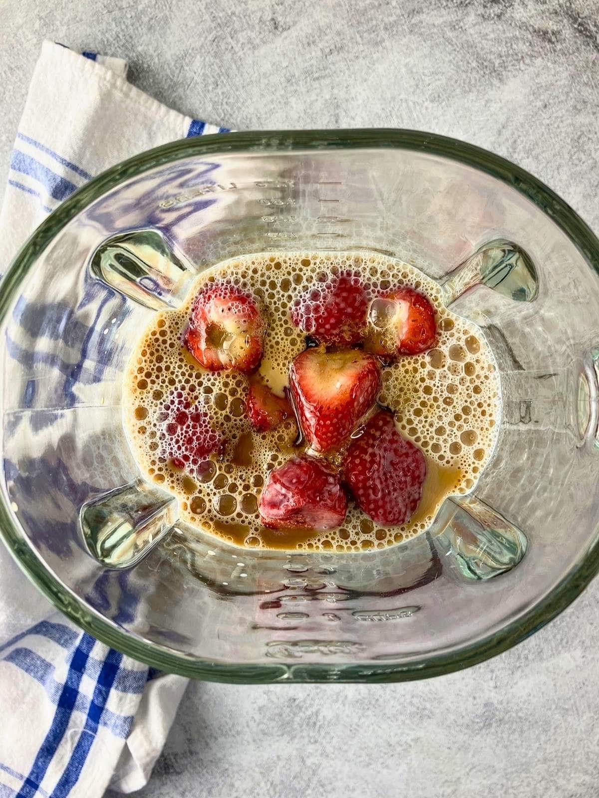 Vanilla and honey added to milk and frozen strawberries in a glass blender before blending. A blue and white towel rests beside the blender on a gray countertop.