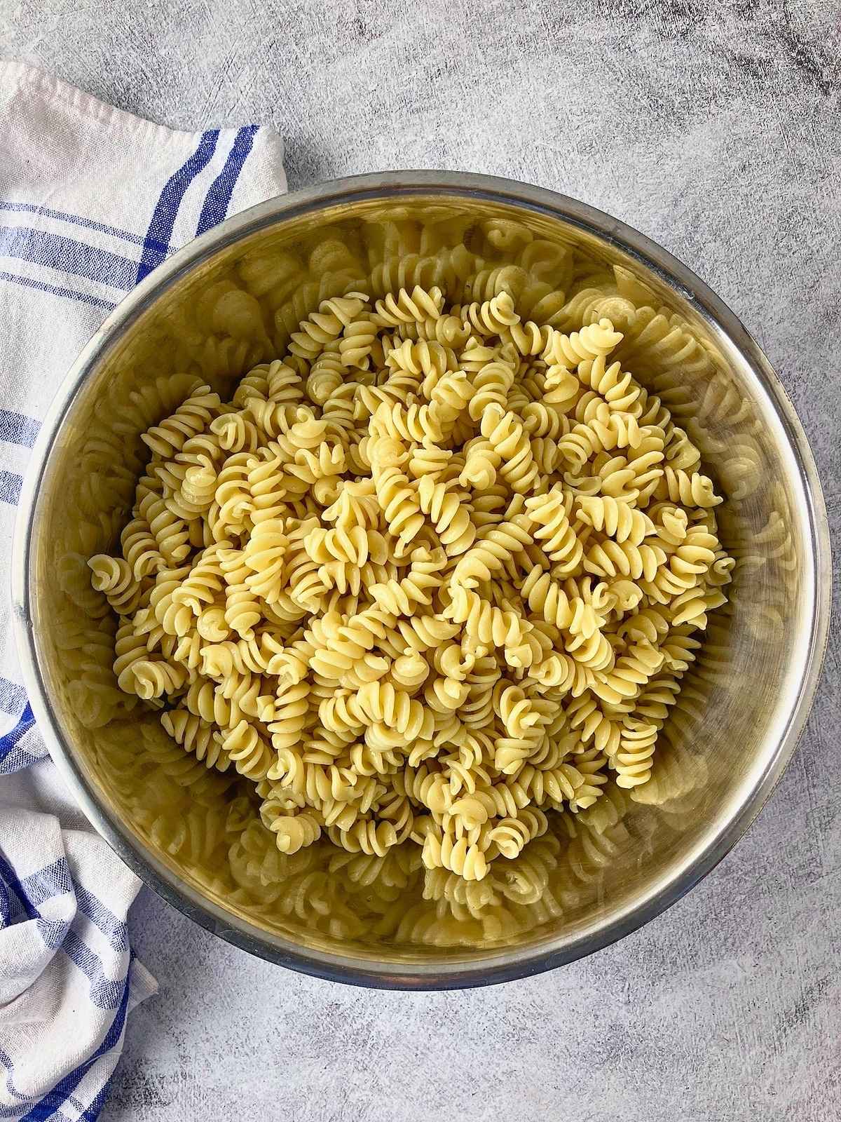 Cooked rotini pasta in a large stainless steel mixing bowl on a gray countertop, ready to be mixed into BLT pasta salad.