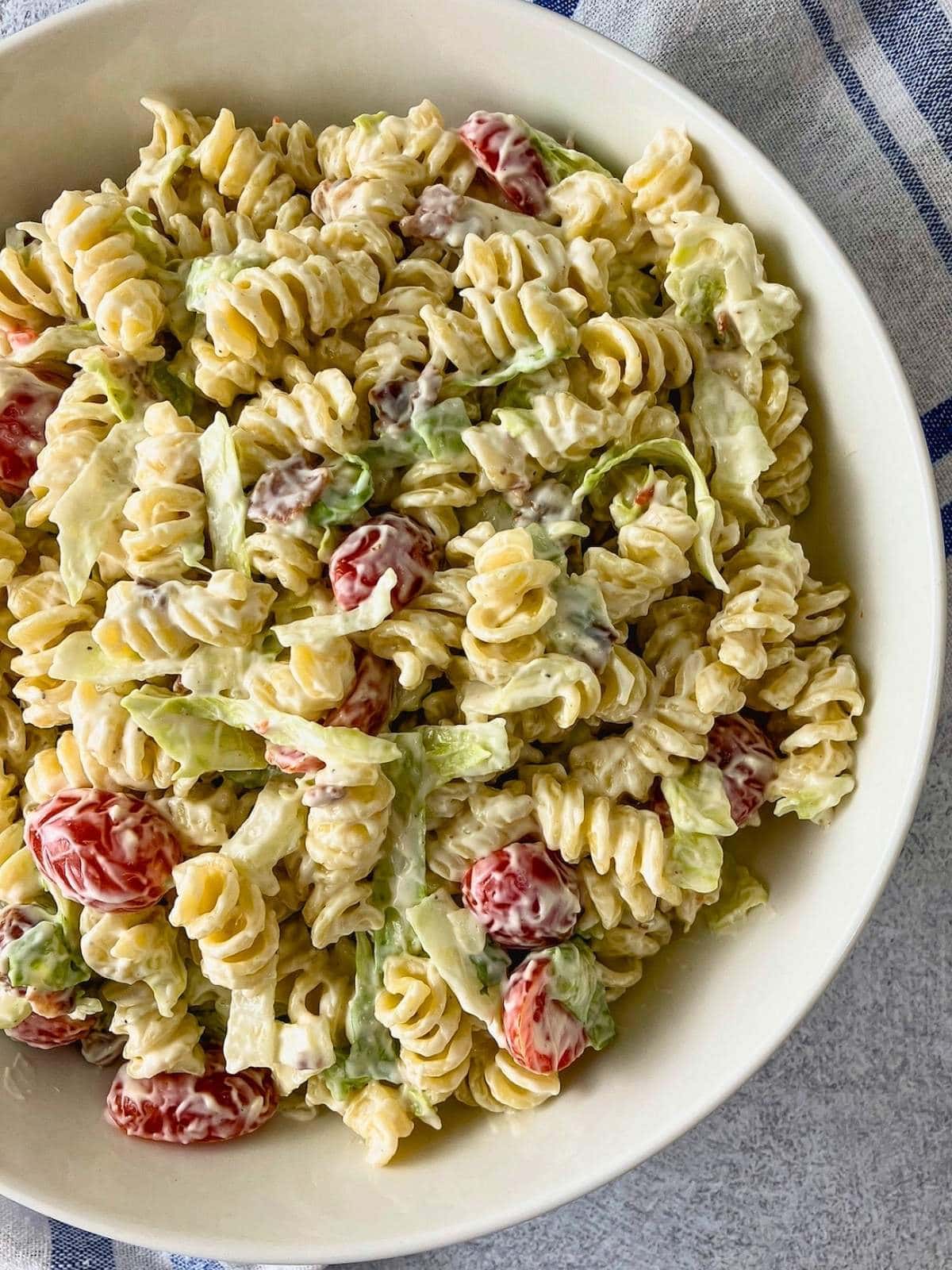 Close up of creamy BLT pasta salad with rotini, grape tomatoes, shredded lettuce, and bacon pieces in a white bowl.