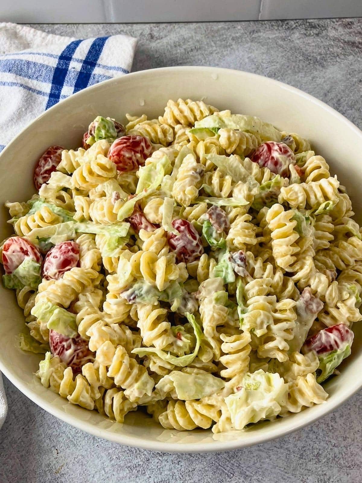 Creamy BLT pasta salad with rotini, grape tomatoes, shredded lettuce, and bacon pieces served in a white bowl beside a blue and white striped towel.