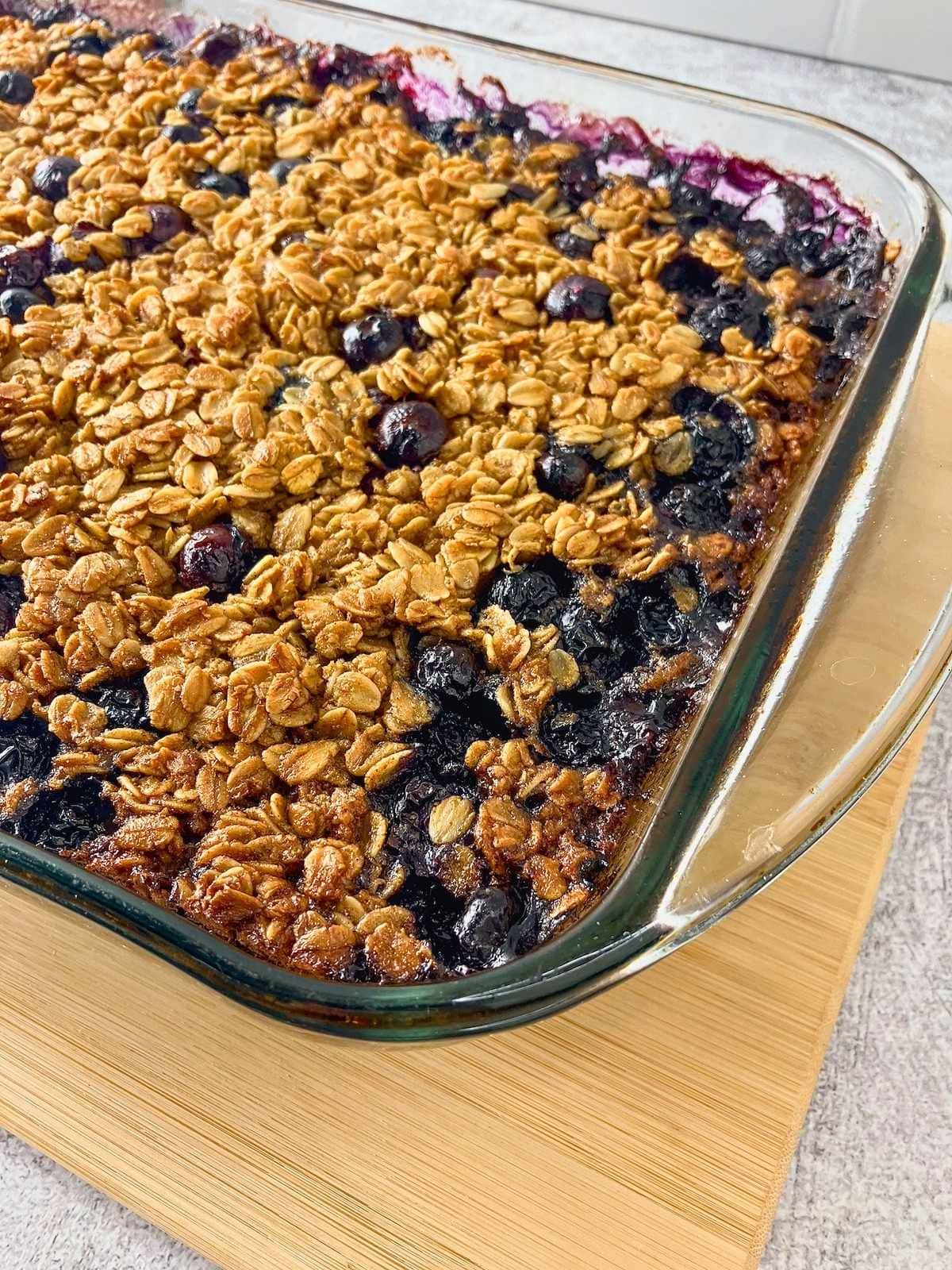 Glass baking dish of blueberry baked oatmeal with golden oats and whole blueberries cooling on a wooden board.