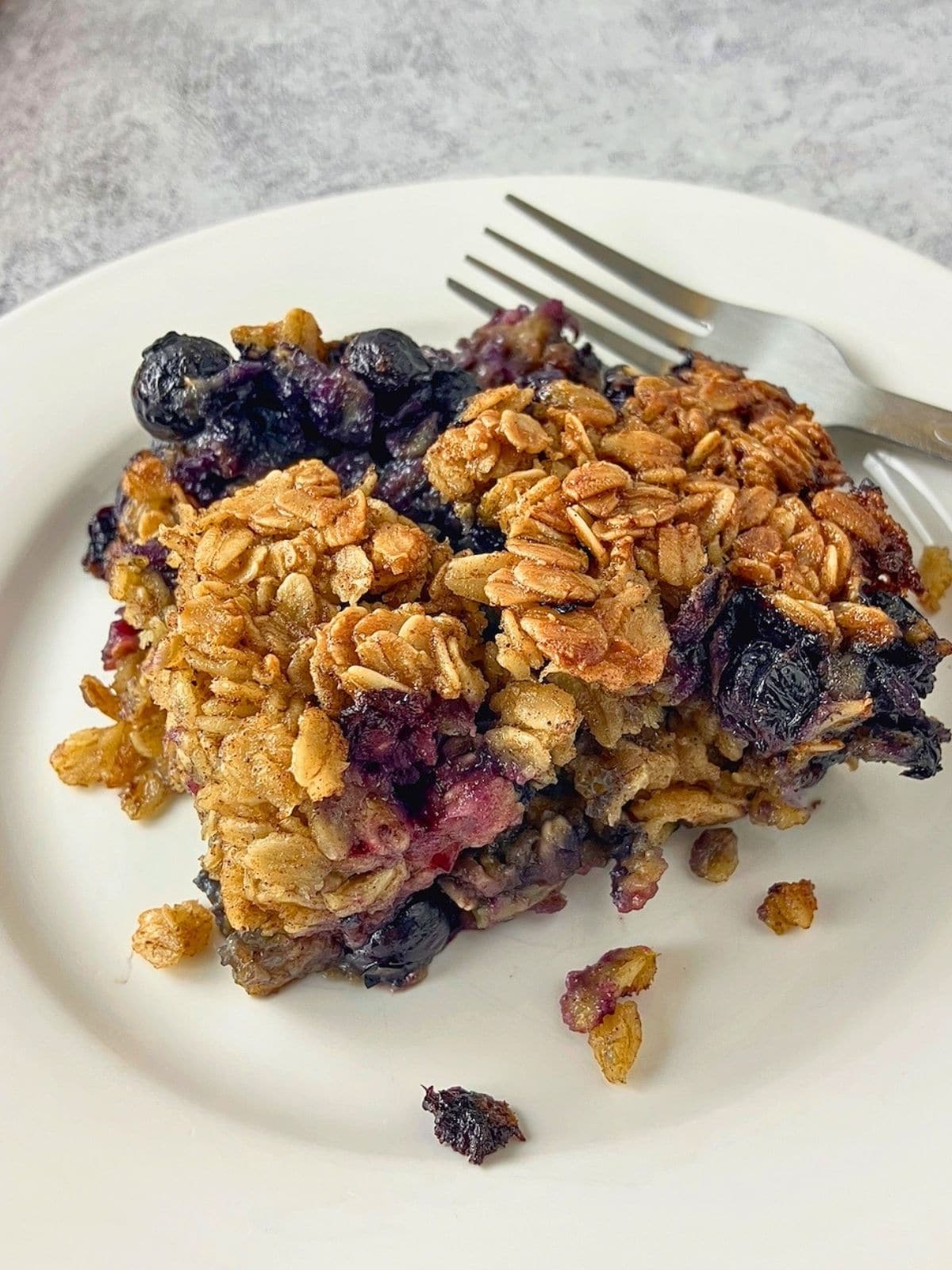 Slice of blueberry baked oatmeal with golden oats and whole blueberries on a white plate with a fork behind it.