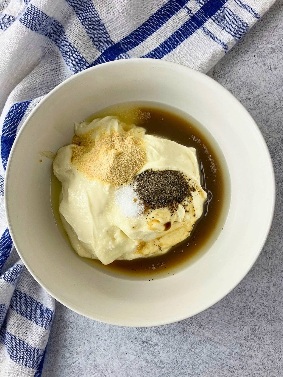 Caesar dressing ingredients in a white bowl with Miracle Whip, Worcestershire sauce, garlic powder, salt, and black pepper before mixing. The bowl sits on a gray countertop beside a blue and white checkered towel.