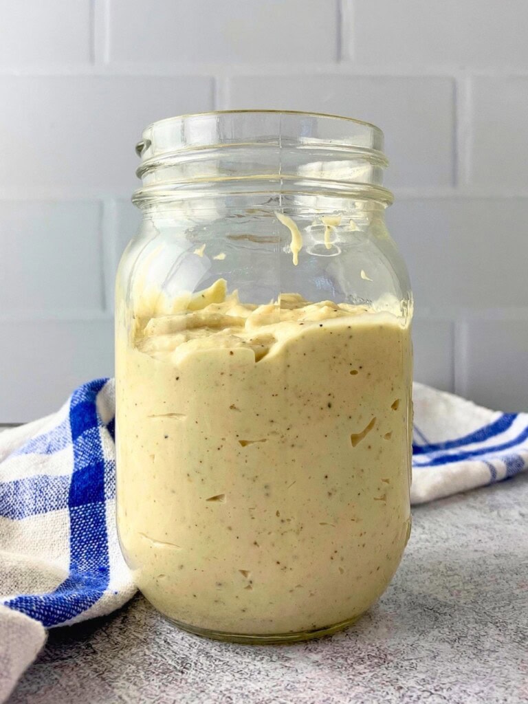 Homemade Caesar dressing in a glass jar on a gray countertop with a blue and white checkered towel behind it. The creamy dressing is thick and speckled with black pepper for a Caesar dressing recipe.