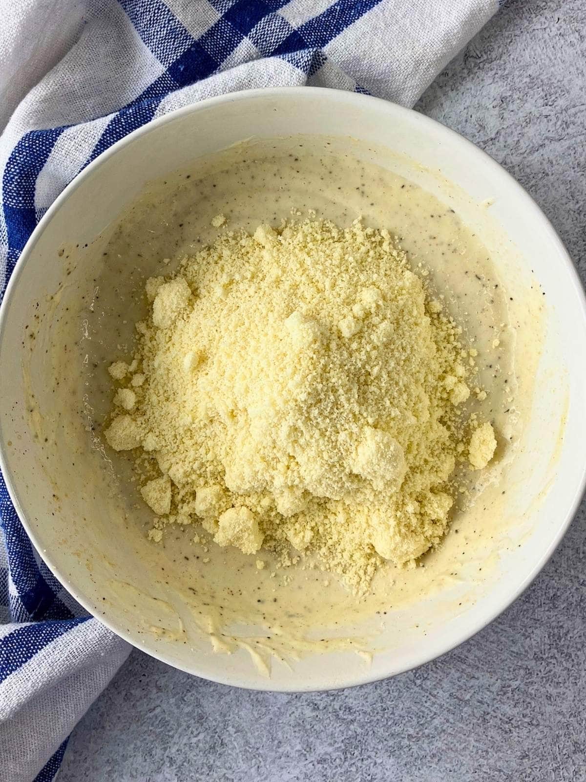 Grated Parmesan cheese added to a bowl of creamy Caesar dressing before mixing. The white bowl sits on a gray countertop beside a blue and white checkered towel.