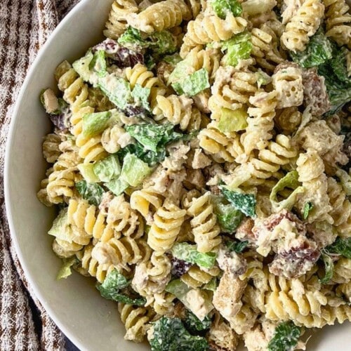 Chicken Caesar pasta salad in a white bowl with rotini, chopped chicken, romaine lettuce, croutons, Parmesan cheese, and creamy dressing. A brown striped kitchen towel sits beside the bowl on a gray countertop.