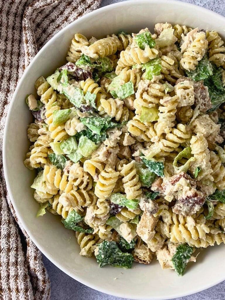 Chicken Caesar pasta salad in a white bowl with rotini, chopped chicken, romaine lettuce, croutons, Parmesan cheese, and creamy dressing. A brown striped kitchen towel sits beside the bowl on a gray countertop.