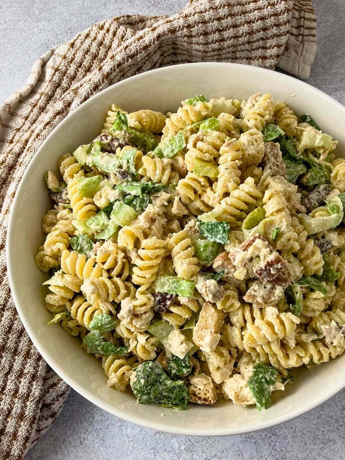 Chicken Caesar pasta salad served in a white bowl with rotini, chopped chicken, romaine lettuce, croutons, Parmesan cheese, and creamy dressing. A brown striped kitchen towel sits behind the bowl on a gray countertop.