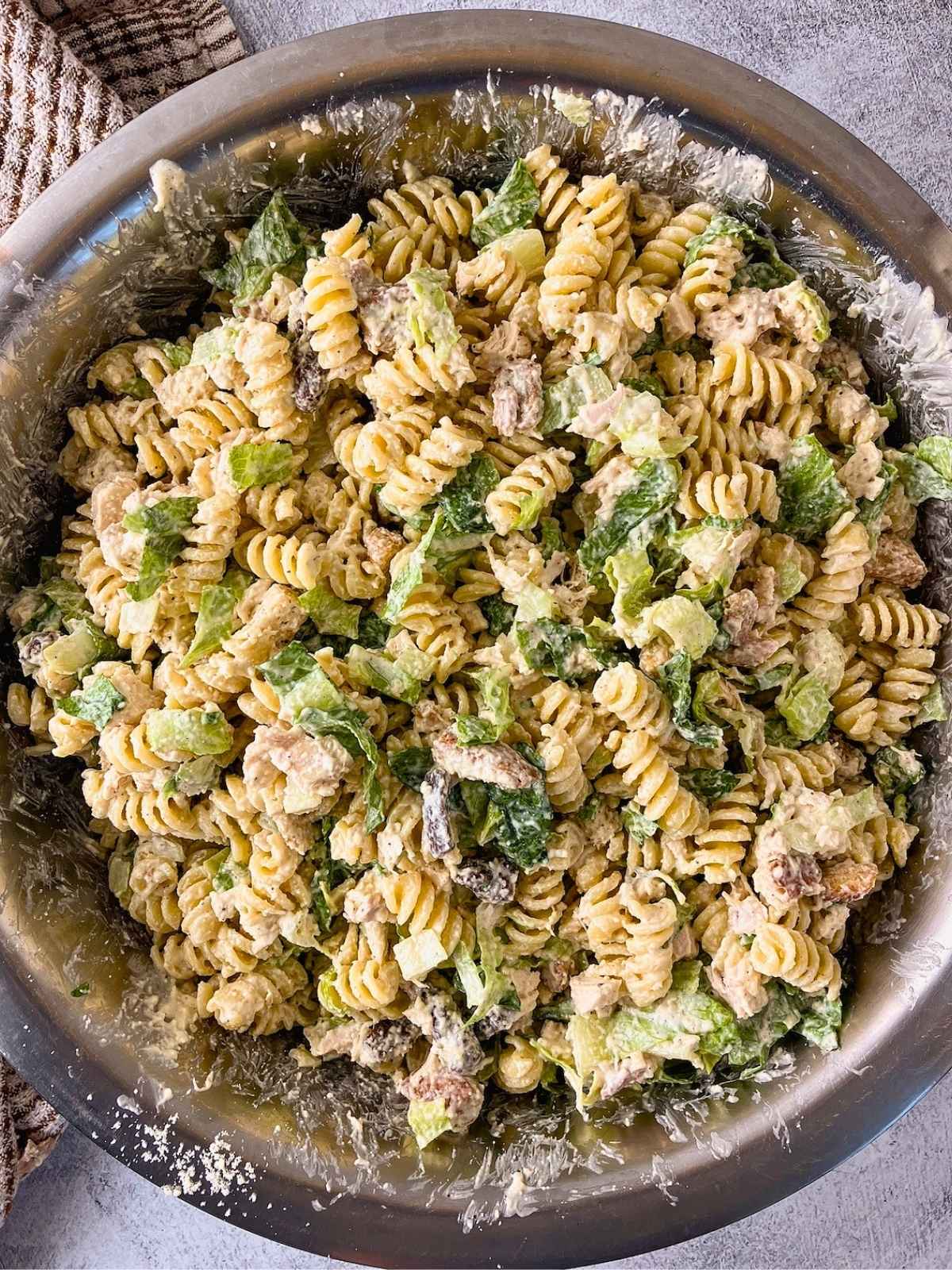 Caesar pasta salad mixed together in a large stainless steel bowl with rotini, chopped chicken, romaine lettuce, croutons, Parmesan cheese, and creamy dressing. A brown striped kitchen towel sits beside the bowl on a gray countertop.